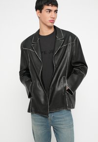 Joven con un blazer negro extragrande de cuero sintético con costuras en contraste, camisa negra y jeans azul claro, mirando hacia un lado.