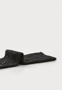 Chaussette noire posée à plat, ornée de taches colorées blanches et vertes, sur un fond blanc uni.