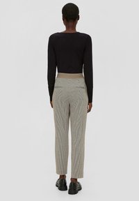 Chemise noire à manches longues avec un col rond, associée à un pantalon beige à motifs avec une taille haute et des poches latérales, porté avec des chaussures noires.