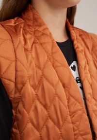 Quilted brezka v bogati oranžni satenski barvi, s šivanim vzorcem v obliki diamanta. Ovratnik je odprt, prekrit s črnim grafičnim majico.