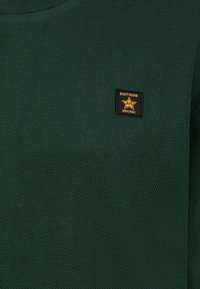 Dunkelgrüner, strukturierter Pullover mit rundem Halsausschnitt und kleinem schwarzem rechteckigem Etikett mit gelbem Text und Stern-Design.