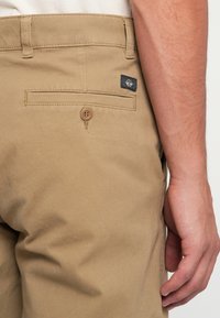 Khaki pants with a smooth, cotton fabric. Features include a back pocket with a button closure and a small black label on the side.