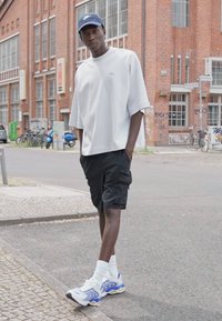 Hvite oversized t-skjorte, svarte cargo shorts, hvite og blå klumpete joggesko, hvite sokker og en blå caps. Urbant bakgrunn med murbygninger.
