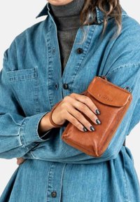 Person i denimkjole holder en brun læderpung med håndleddet, har sorte malede negle og bærer en mørkegrå turtleneck under.