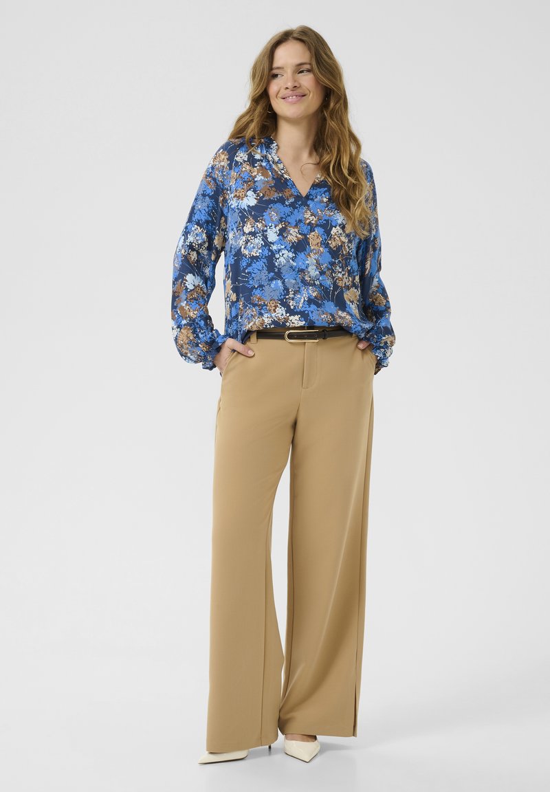 Blommig blå blus med långa ärmar och V-ringning, matchad med beige vida byxor och ett svart bälte. Vita klackskor fulländar outfiten.