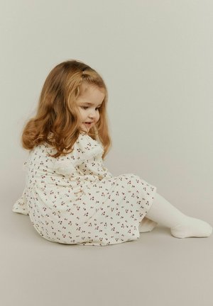 White dress with small red and brown floral patterns, ruffled neckline, and soft texture. Paired with white tights. Child sitting sideways.