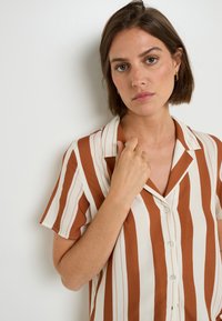 Camicia a maniche corte a righe marroni e crema con colletto, dotata di chiusura con bottoni e una texture del tessuto liscia e leggera.