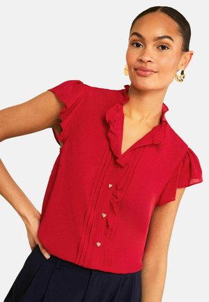 Frau trägt eine rote ärmellose Bluse mit Rüschen und herzförmigen Knöpfen sowie goldene Creolen, posiert mit der Hand in der Hüfte vor weißem Hintergrund.