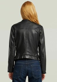 Persona bionda che indossa una giacca di pelle nera aderente e jeans blu, in piedi con le spalle rivolte verso un fondo chiaro e semplice.