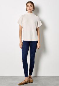 Helles beige Top mit kurzen Ärmeln und hohem Kragen, gerippte Textur. Kombiniert mit dunkelblauen, eng anliegenden Jeans und braunen Wildlederschuhen. Minimalistisches Design.