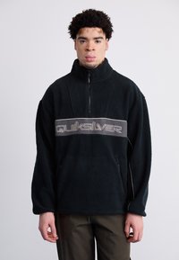 Sort fleece-pullover med høj krave, der har et beige Quiksilver-logo på brystet og lynlåslommer på siden. Blød tekstur.