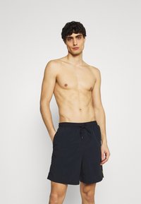 Pantaloni da mare blu navy con cintura elastica e cordino, tasche laterali e un logo sul lato inferiore della gamba. Tessuto liscio e leggero.