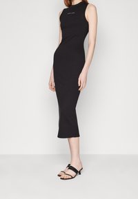 Svart ärmlös midi-klänning med en figurnära silhuett och sidoklyfta. Har en rund halsringning och subtil logotypdetalj vid bröstet.