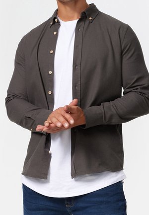 Mann trägt ein aufgeknöpftes dunkelgraues Langarmhemd über einem weißen T-Shirt und blauen Jeans, die Hände vor dem Körper verschränkt.