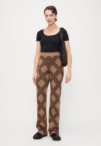 Bruine broek met een patroon van zwarte geometrische ontwerpen, gecombineerd met een zwart aansluitend topje en een zwarte schoudertas. Platte zwarte schoenen.