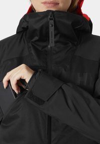 Czarna wodoodporna kurtka z wysokim kołnierzem, kapturem i kieszeniami na zamek. Posiada rzep na rękawie oraz logo nadrukowane na piersi.