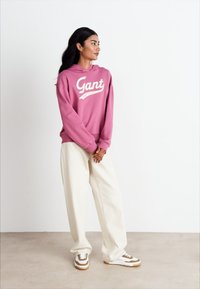 Pink hoodie with "Gant" logo, white lettering, loose fit. Paired with cream wide-leg pants and white sneakers with brown accents.