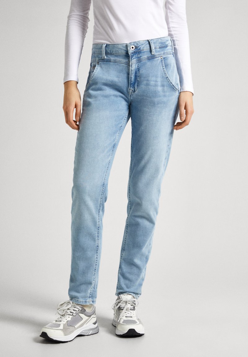 Jeans de denim azul claro con un ajuste slim, que cuentan con un cierre de botón, bolsillos frontales y una textura ligeramente desgastada. Llevados con zapatillas grises y blancas.