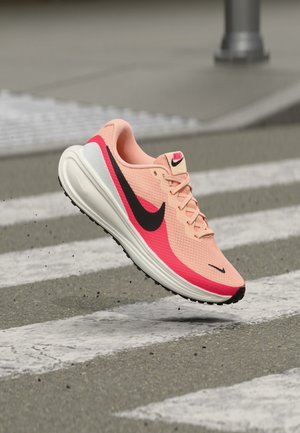 Chaussure de course Nike rose clair et rouge en l'air au-dessus d'un passage piéton sur une route en asphalt.