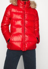 Rød puffjakke med skinnende, quiltet stof, med en pelsforet hætte, lynlås foran og sidelommer, parret med mørke jeans.