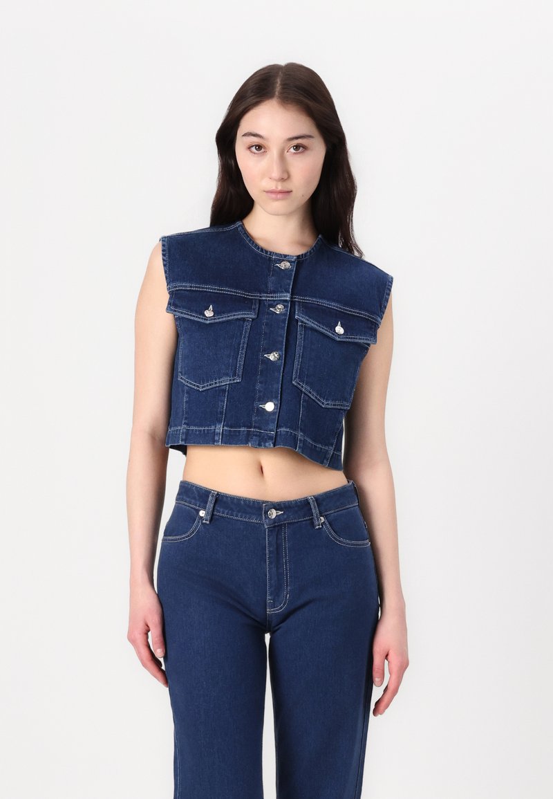 Gilet in denim, cropped e senza maniche, di un blu scuro, con chiusura a bottoni, due tasche sul petto e scollatura rotonda, abbinato a pantaloni in denim.
