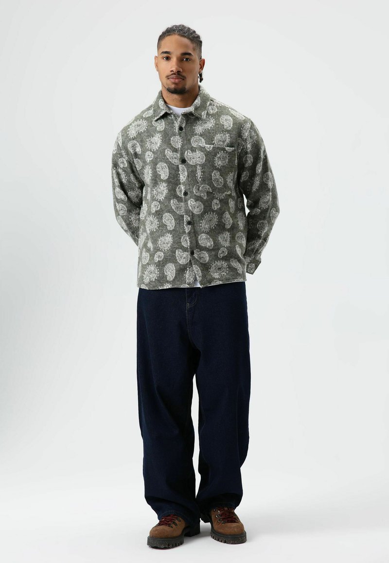 Grünes Hemd mit Muster und Brusttasche, kombiniert mit locker sitzenden dunkelblauen Jeans und braunen Schuhen. Neutraler Hintergrund.