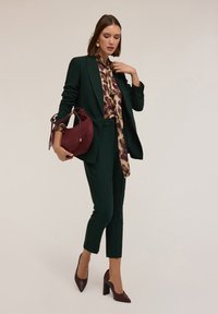 Completo su misura verde composto da pantaloni e giacca, abbinato a una blusa a fantasia e una borsa bordeaux. Il modello indossa scarpe con tacco bordeaux.