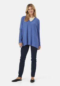 Suéter azul de cuello en V hecho de una tela suave, con mangas largas y un corte holgado, combinado con pantalones vaqueros oscuros y zapatos negros.
