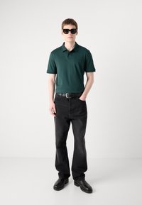 Tmavozelená polo košeľa, čierne rifle a čierne topánky. Model nosí nadrozmerné čierne slnečné okuliare a stojí na jednotnom pozadí.