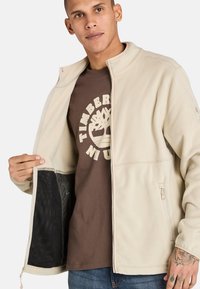 Beige fleeceställning med hög krage, dragkedja och fickor. Bärs över en brun T-shirt med en trädlogotyp. Strukturerad tyg.