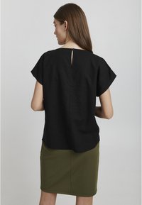 Top corto nero a maniche corte con una vestibilità ampia, caratterizzato da una chiusura a chiave sul retro. Indossato con una gonna a tubino verde, che mette in risalto il tessuto strutturato.
