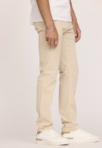 Pantalons en coton beige à coupe slim, présentant une texture lisse et des détails de coutures courbées, associés à des baskets blanches.