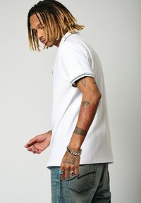 Polo shirt blanc en tissu texturé avec des poignets à motifs, associé à un jean bleu clair. Présente plusieurs bagues et un bracelet en argent.