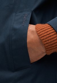 Μπλε ναυτικό μπουφάν με υφασμάτινη υφή, που διαθέτει ορατό λογότυπο Barbour στην τσέπη, σε συνδυασμό με ριμπ πλεκτή μανσέτα πουλόβερ σε σκουριασμένο χρώμα.