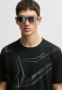 Black T-shirt with a geometric silver pattern across the front, short sleeves, and a round neckline. Black mirrored sunglasses complete the look.