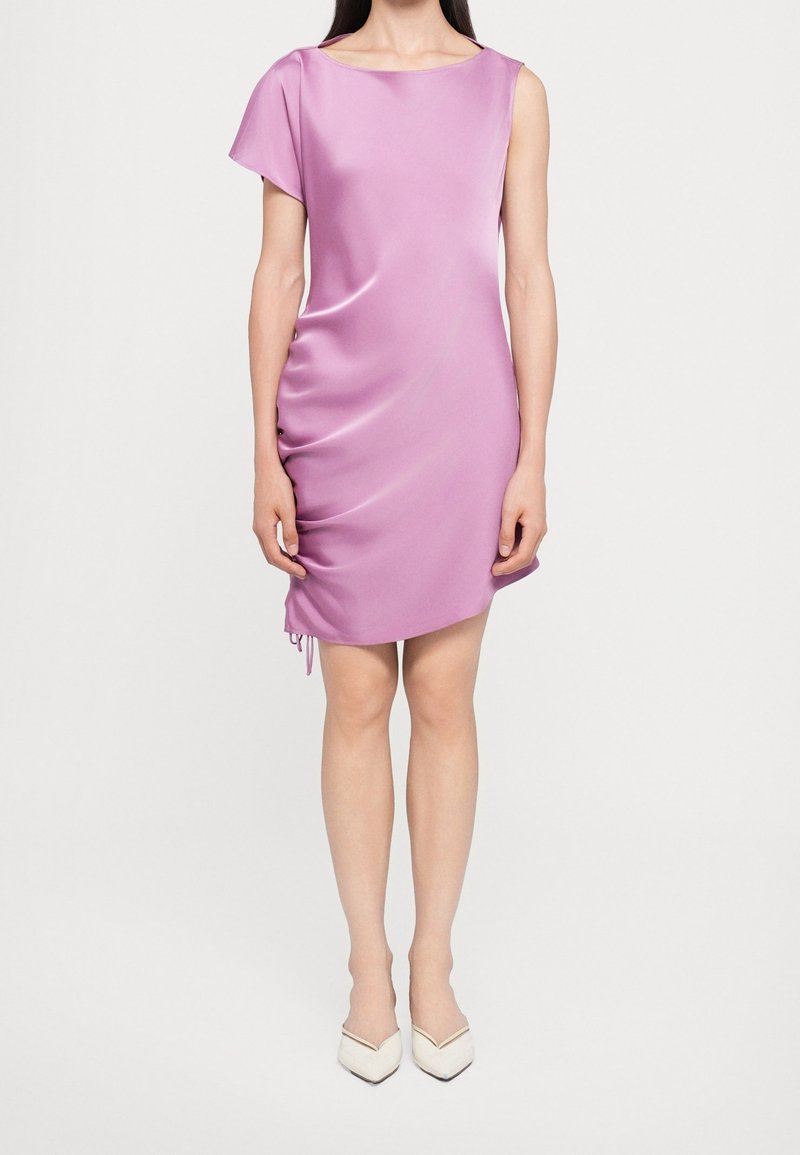Femme portant une robe courte en satin rose brillant avec une manche courte et des talons blancs à bout pointu, debout sur un fond blanc.