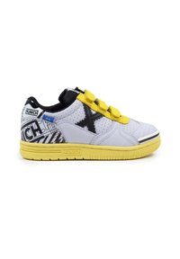 Zapatilla blanca de corte bajo con logo negro en forma de "X", suela y tiras amarillas, talón gráfico en blanco y negro, y collar acolchado negro.