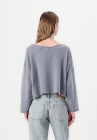 Suéter cinza tricotado, cortado, com um decote largo e bordas desfiadas, combinado com jeans de cintura alta em azul claro, exibindo um ajuste descontraído.