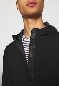Nahaufnahme einer Person, die eine schwarze Kapuzenjacke über einem schwarz-weiß gestreiften Hemd trägt, wobei der Reißverschluss der Jacke teilweise geöffnet ist.