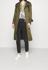 Oliivinvihreä trenssitakki kaksirivisillä napeilla, yhdistetty kuvioituun paitaan, mustiin farkkuihin ja valkoisiin tennareihin. Sileä pinta.