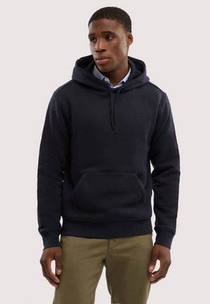 Mann trägt eine marineblaue Kapuzenjacke über einem hellblauen Hemd mit Kragen und khakifarbene Hose, schaut leicht nach links, die Hände entspannt an den Seiten.