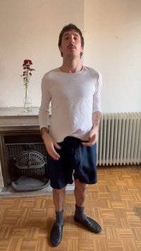 Vyras stovintis patalpoje prie židinio ir radiatoriaus, dėvintis baltą ilgomis rankovėmis marškinį, tamsiai mėlynas trumpąsias kelnes, pilkas kojines ir juodus šlepetes.