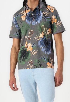Homme portant un polo gris avec des imprimés floraux tropicaux bleus, verts et orange, et un jean bleu clair sur fond blanc.