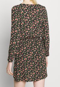 Robe fleurie à manches longues, en tissu vert foncé, ornée de petites fleurs roses et jaunes. Taille élastique et encolure ronde avec un détail de bouton.