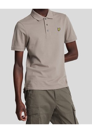 Homme portant un polo beige à manches courtes avec trois boutons et un logo jaune, associé à un pantalon cargo vert olive.