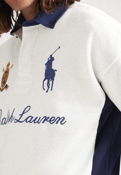 Λευκό πόλο πουκάμισο με σκούρο μπλε γιακά, σκούρο μπλε λογότυπο Ralph Lauren με παίκτη πόλο πάνω σε άλογο και κειμήλιο κεντημένο με τη λέξη «Ralph Lauren» στο στήθος.