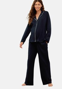 Smiling woman wearing black long-sleeve buttoned pajamas with white trim, standing barefoot against a plain background.