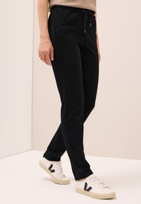 Person in schwarzen Jogginghosen mit Kordelzug und weißen Sneakers mit schwarzen "V"-Logos, steht vor einer hellbeigen Wand und einem hellbeigen Boden.