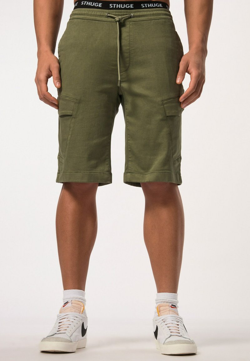 Shorts cargo vert olive avec deux poches latérales, ceinture élastique et cordon de serrage. Associés à des baskets blanches et des chaussettes visibles.