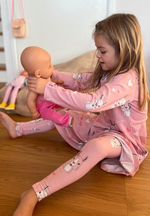 Kind, das einen pinken Schlafanzug mit Hasen- und Herzmustern trägt und eine Puppe hält. Der Hintergrund zeigt einen Holzboden und eine weiche beige Wand.